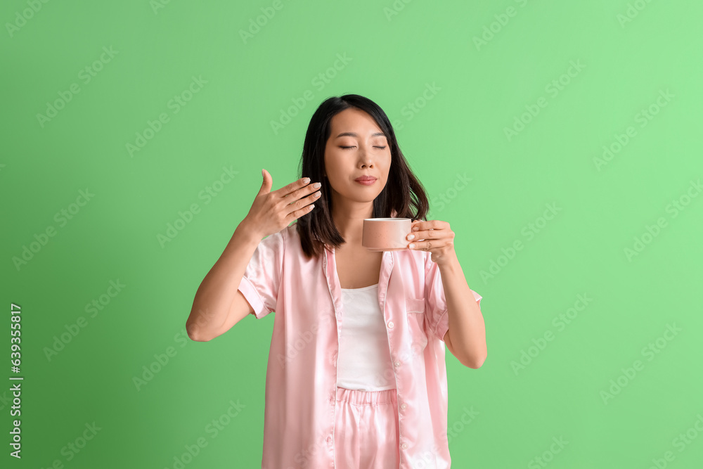 Beautiful Asian woman in pajamas with cup of coffee on green background