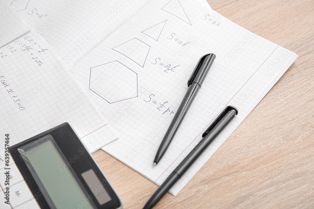 Open copybooks with math formulas, calculator and pens on wooden table, closeup