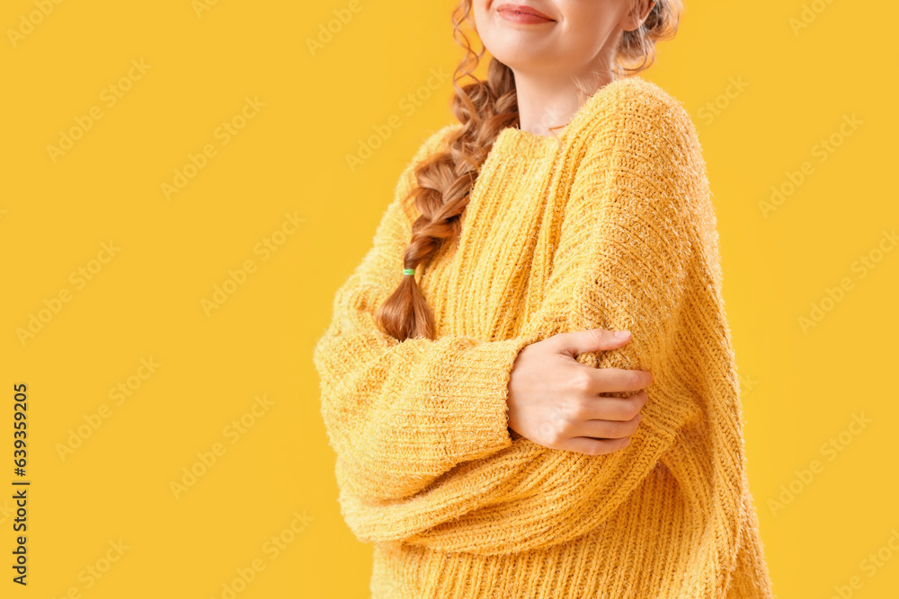 Young woman in warm sweater and eyeglasses hugging herself on yellow background