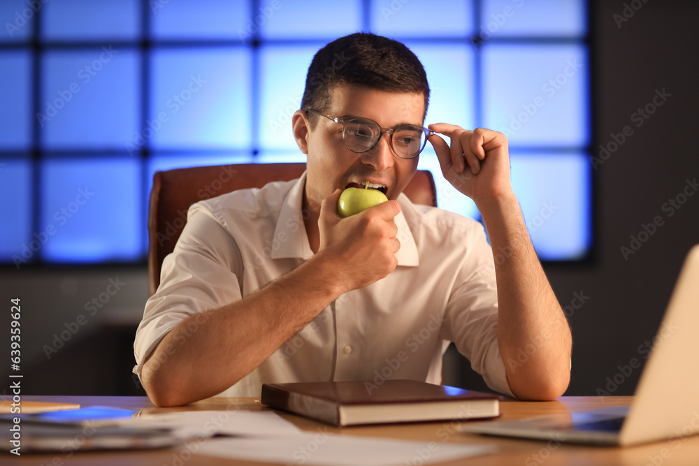 Young businessman eating apple in office at night