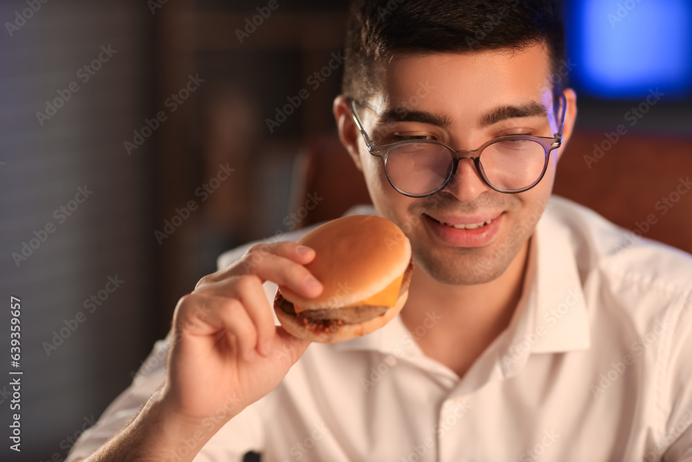 Young businessman with tasty burger in office at night, closeup