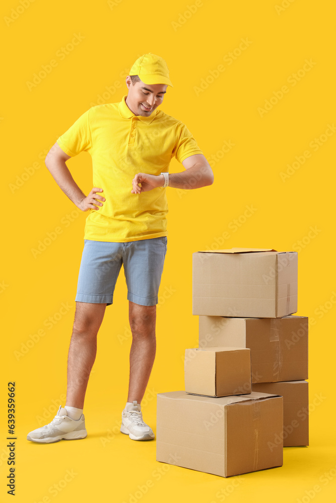 Male courier with parcels looking at smartwatch on yellow background