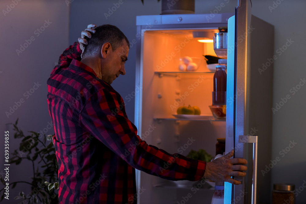 Hungry mature man near open fridge in kitchen at night