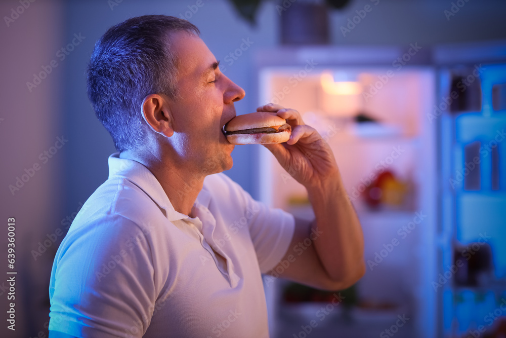 Hungry mature man eating burger in kitchen at night, closeup