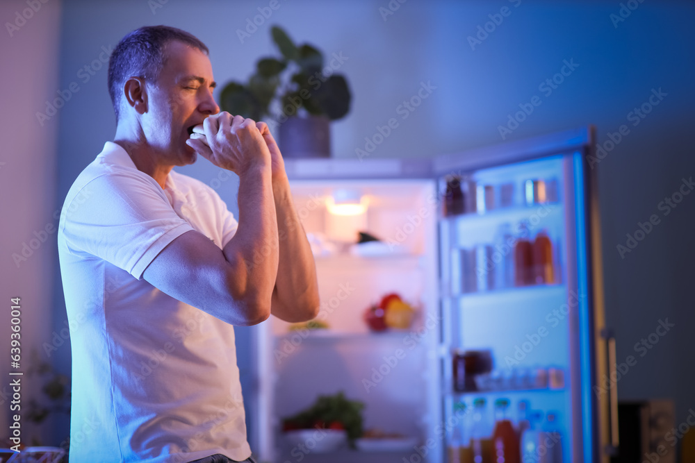 Hungry mature man eating burger in kitchen at night