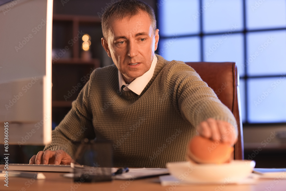 Mature man eating burger in office at night