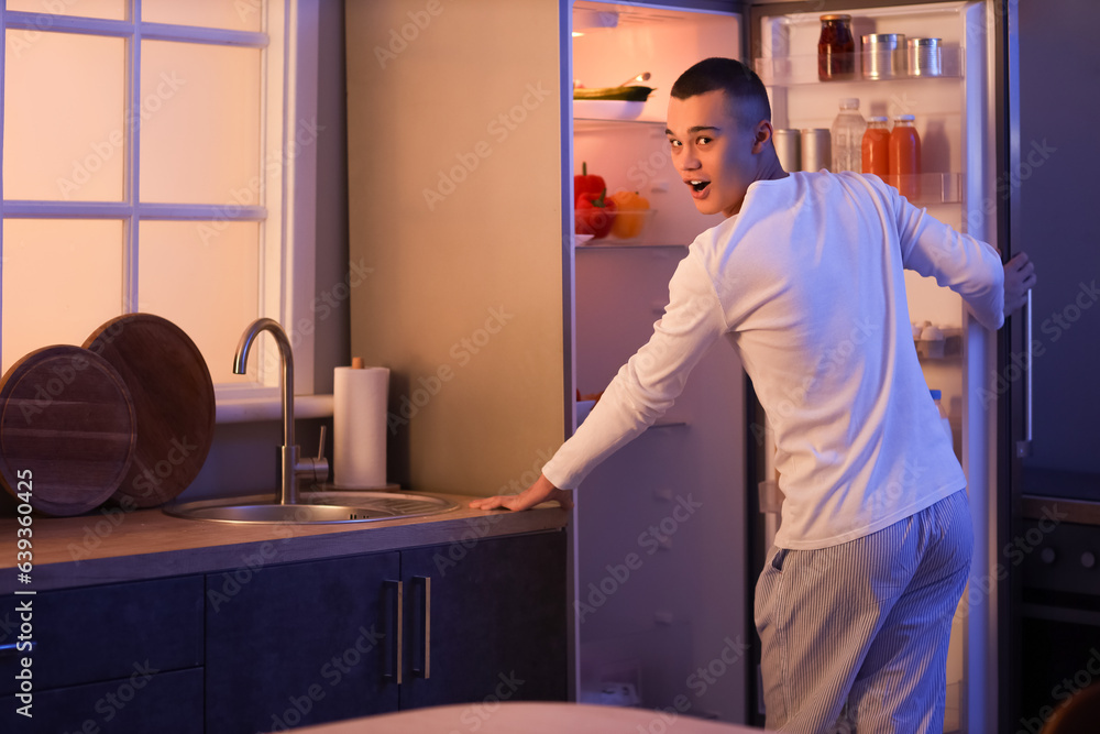 Hungry young man near open fridge in kitchen at night