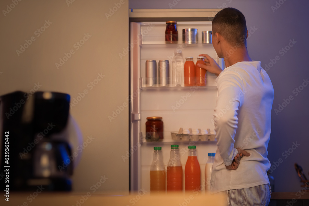 Hungry young man near open fridge in kitchen at night, back view