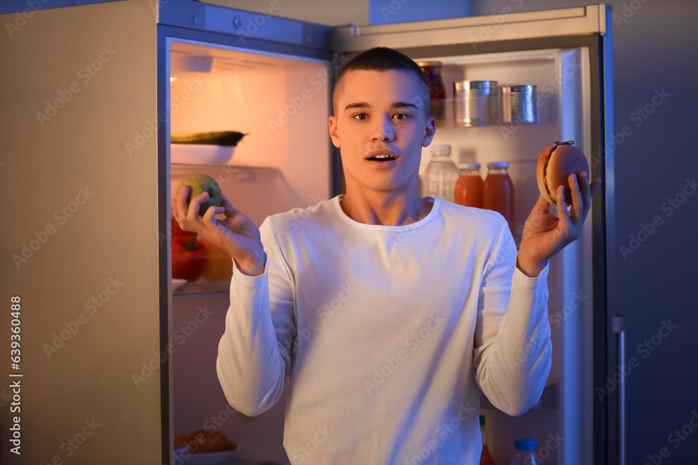 Hungry young man with apple and burger near open fridge in kitchen at night