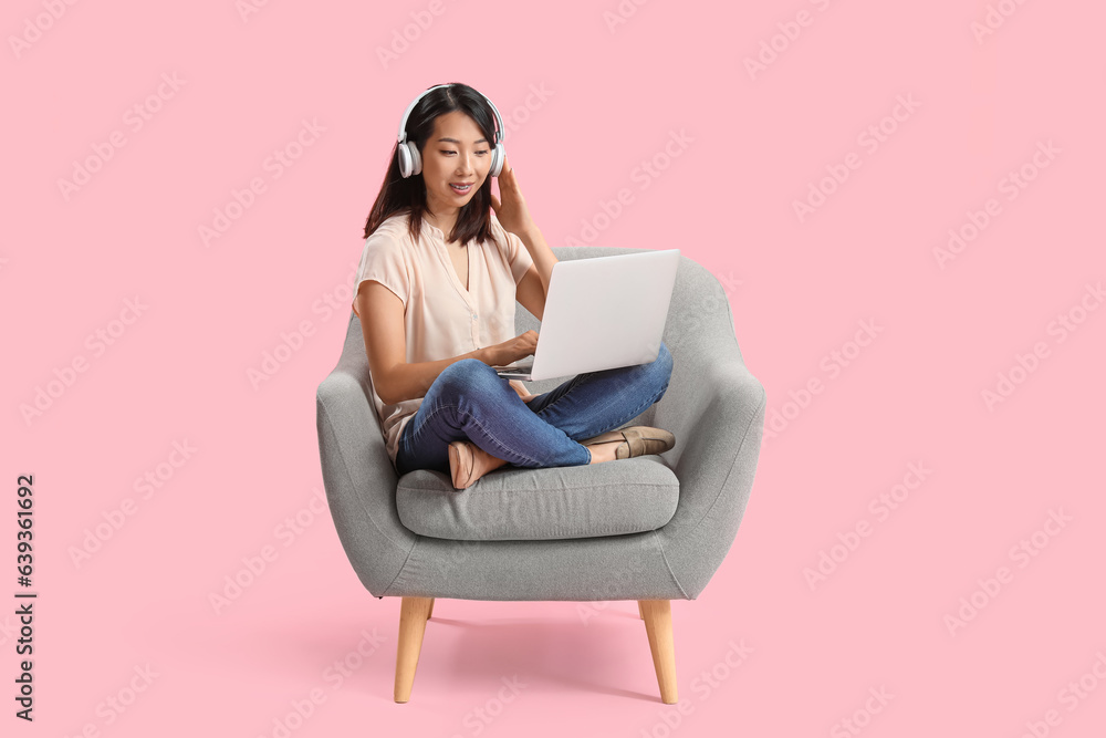 Beautiful Asian woman with headphones using laptop in grey armchair on pink background