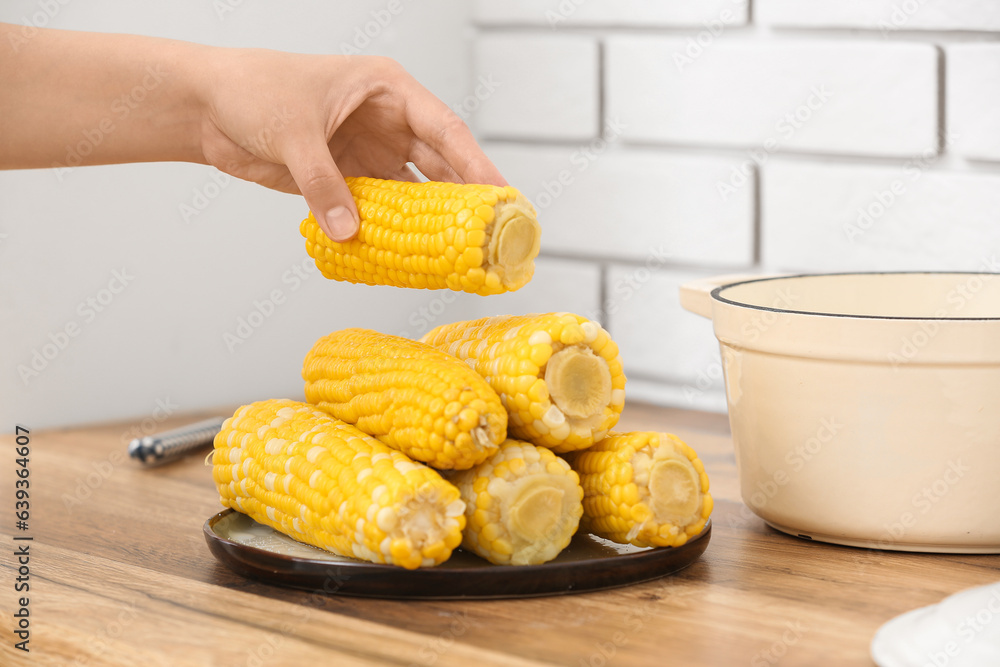 Woman taking boiled corn cobs from plate on wooden table in kitchen