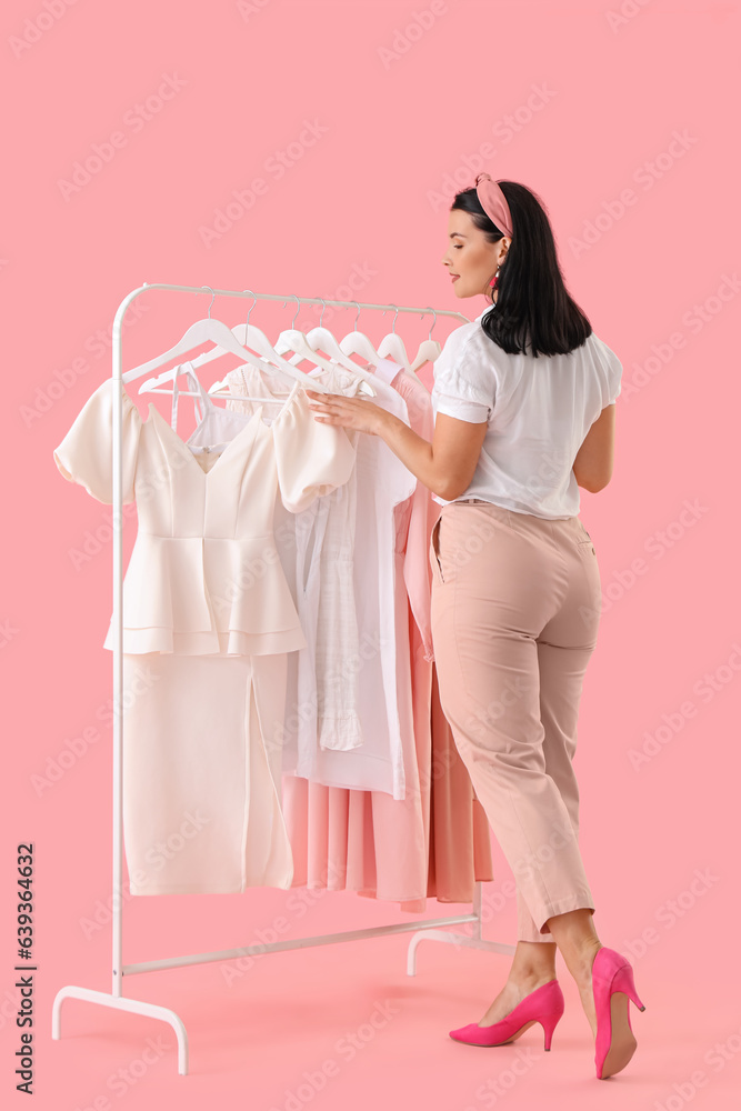 Beautiful young woman choosing clothes on pink background