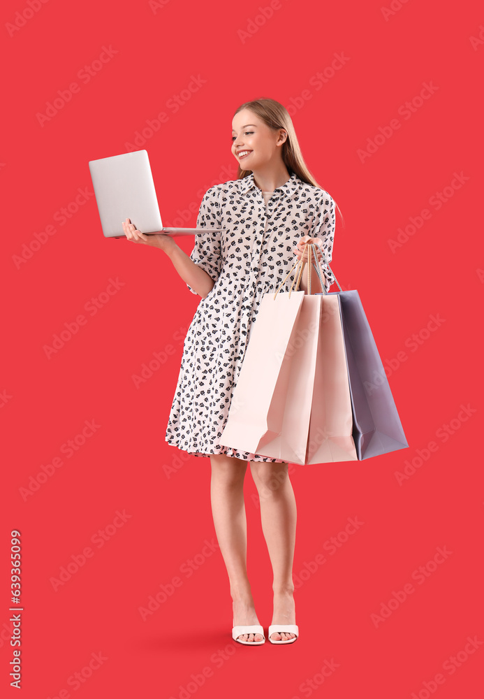 Pretty young woman with laptop and shopping bags on red background. Online shopping concept