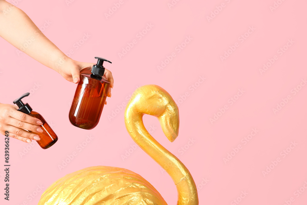 Female hands holding cosmetic products with decorative flamingo on pink background