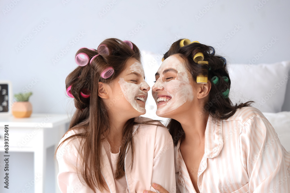 Little girl and her mother with hair curlers applying facial mask in bedroom, closeup