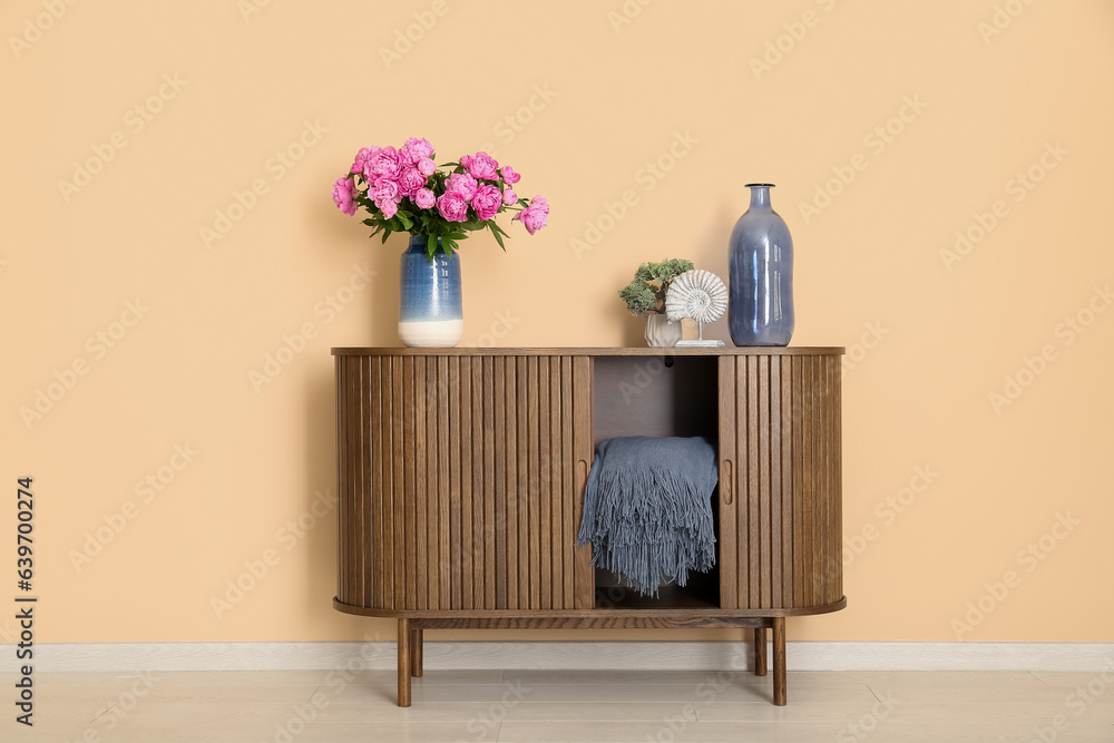 Vase of pink peonies with figurine and houseplant on dresser near beige wall