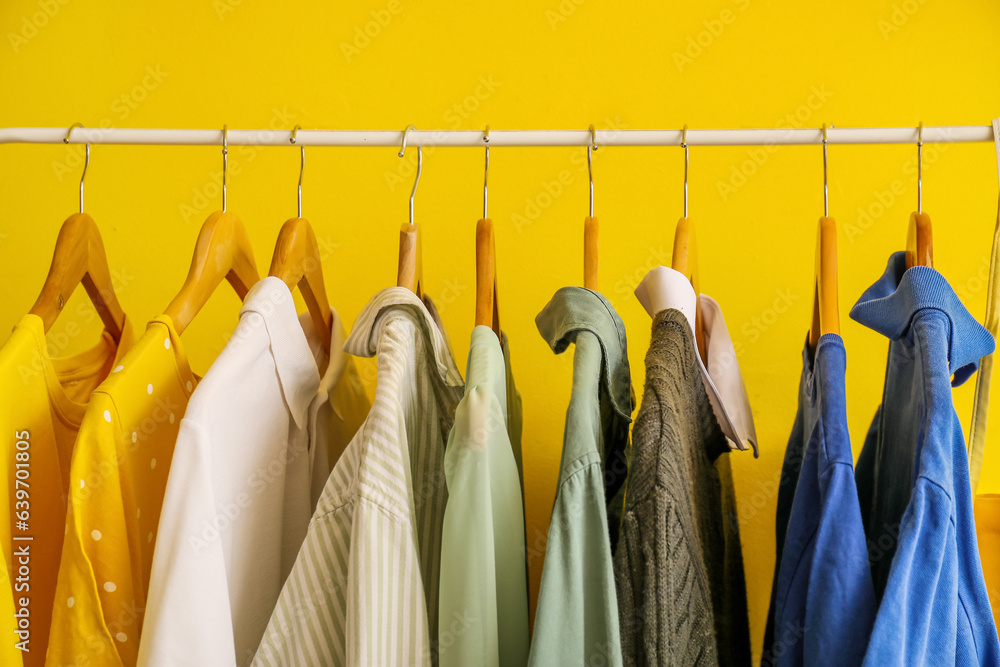 Different clothes hanging on rack against yellow background, closeup