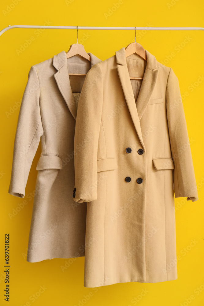 Stylish coats hanging on rack against yellow background, closeup