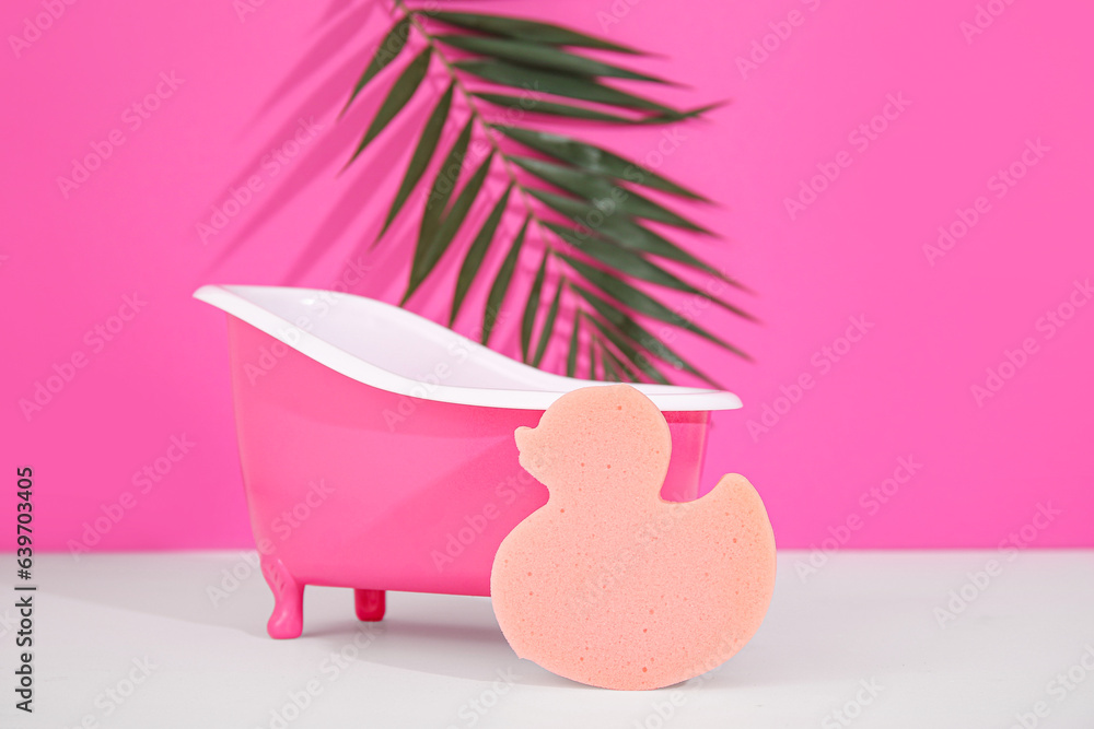 Small bathtub, sponge and palm leaf on light table against color background