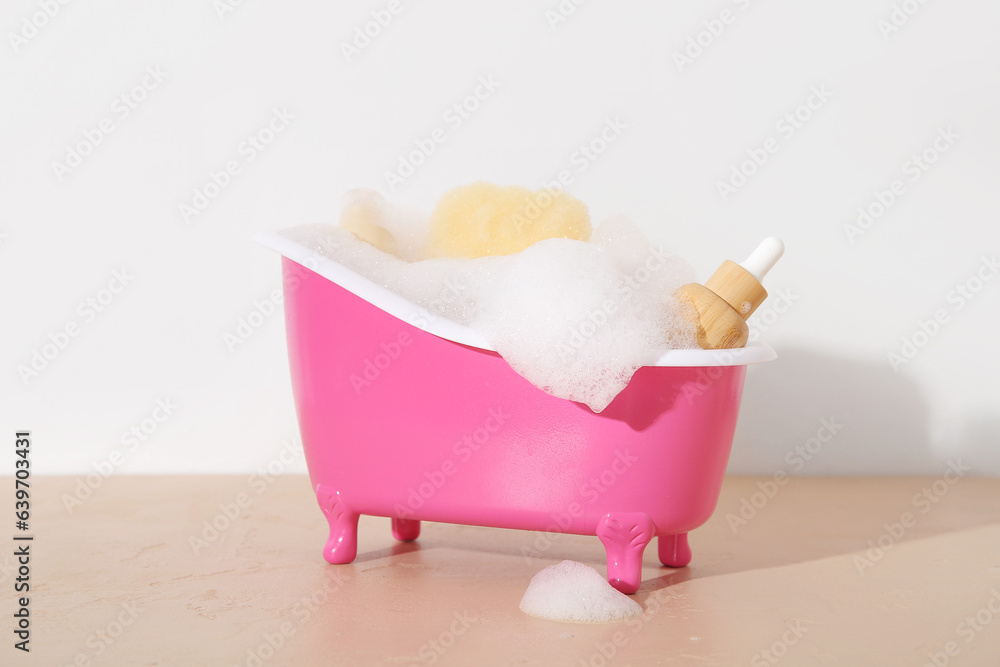 Small bathtub with foam, cosmetic products and sponge on color table against light background