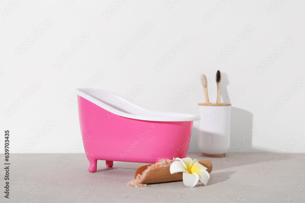 Small bathtub, sea salt and toothbrushes on table against light background