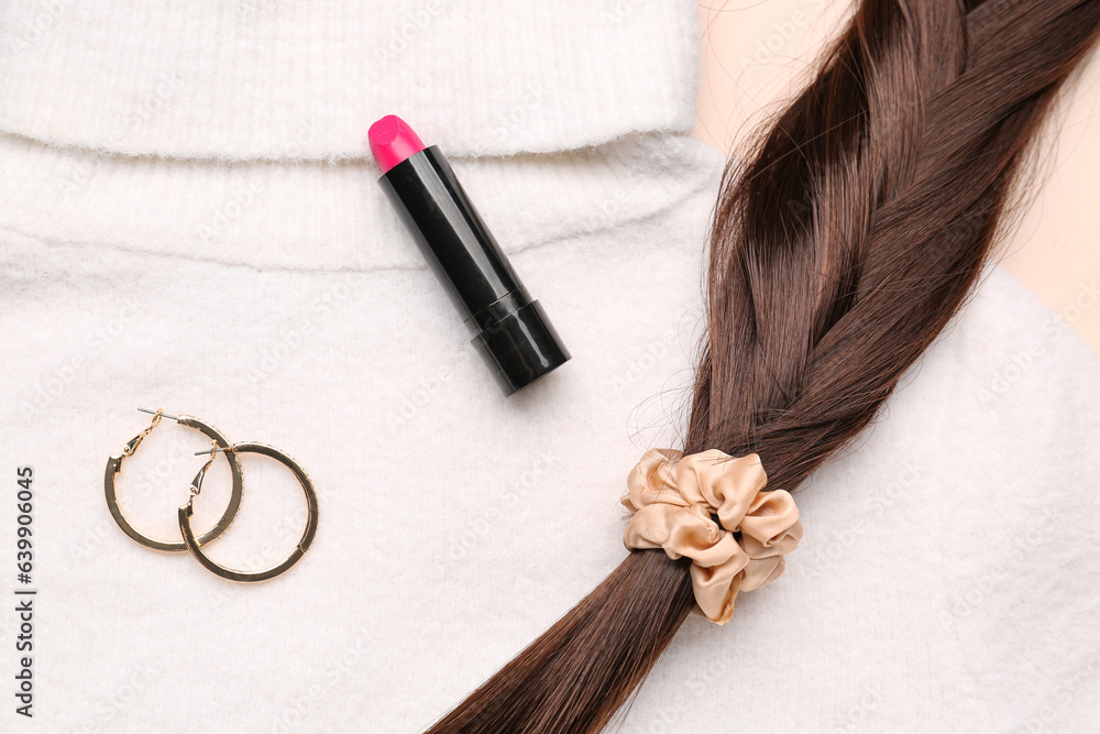 Braided hair strand with scrunchy, lipstick, earrings and sweater on beige background, closeup