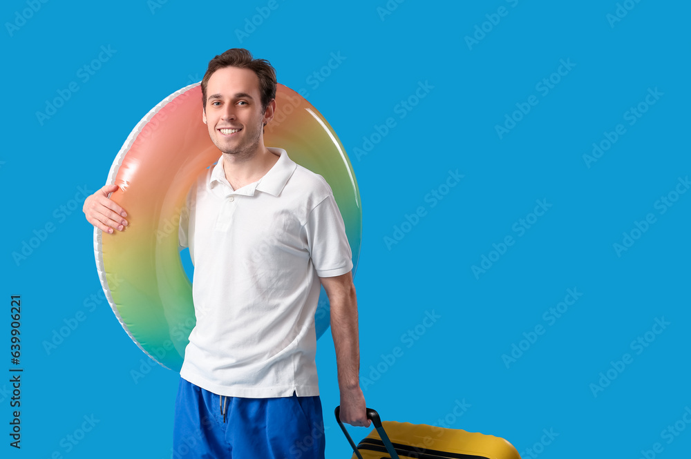 Young man with inflatable ring and suitcase on blue background