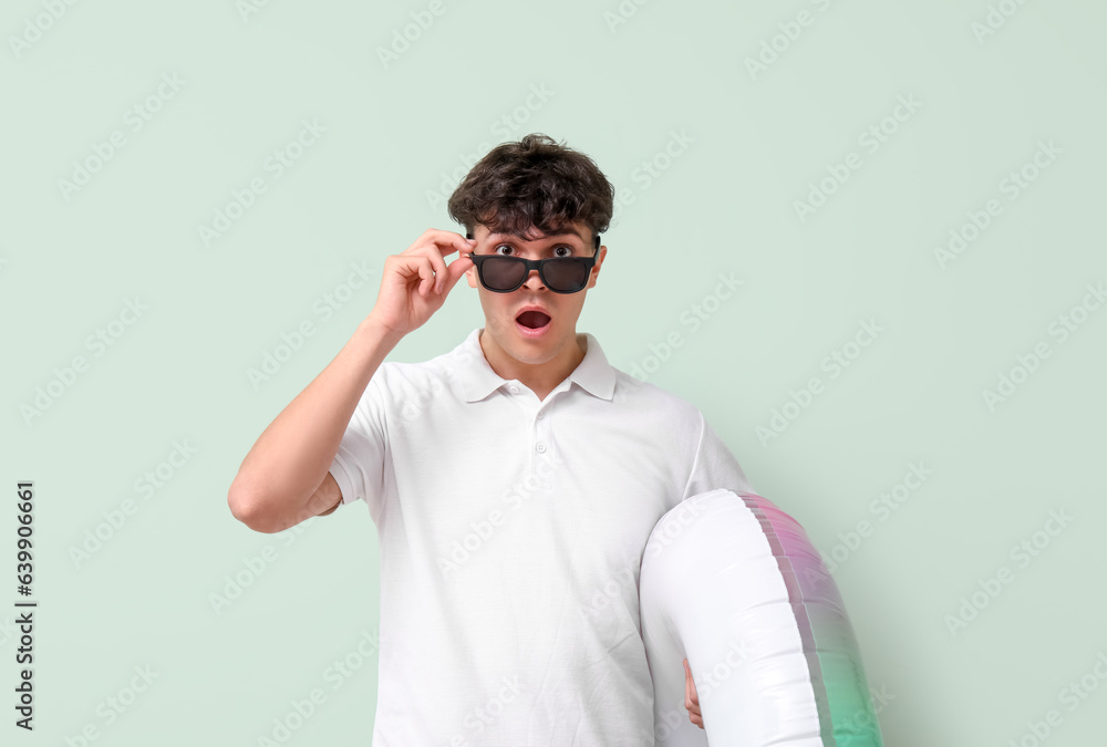 Shocked young man with sunglasses and inflatable ring on green background