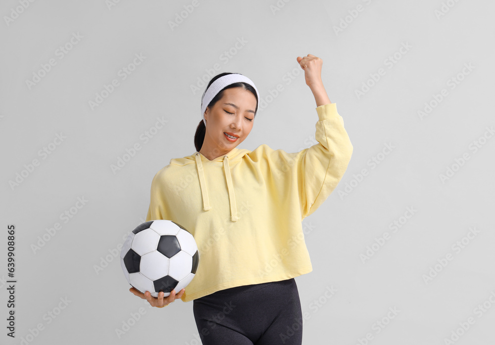 Happy young sporty Asian woman with soccer ball on grey background