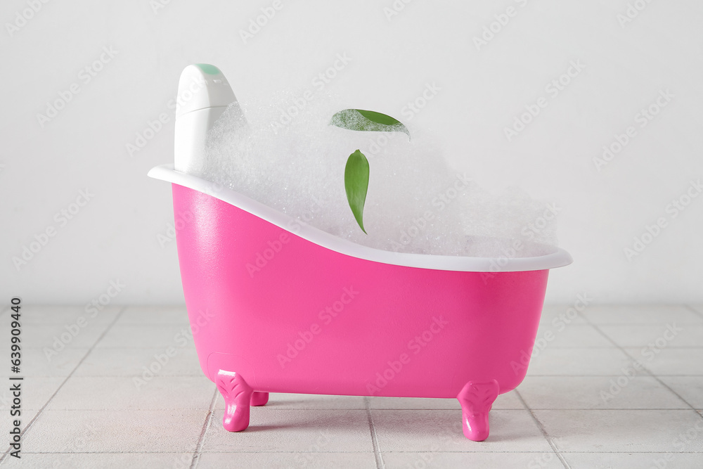 Small bathtub with foam and bottle of cosmetic product on light tile table