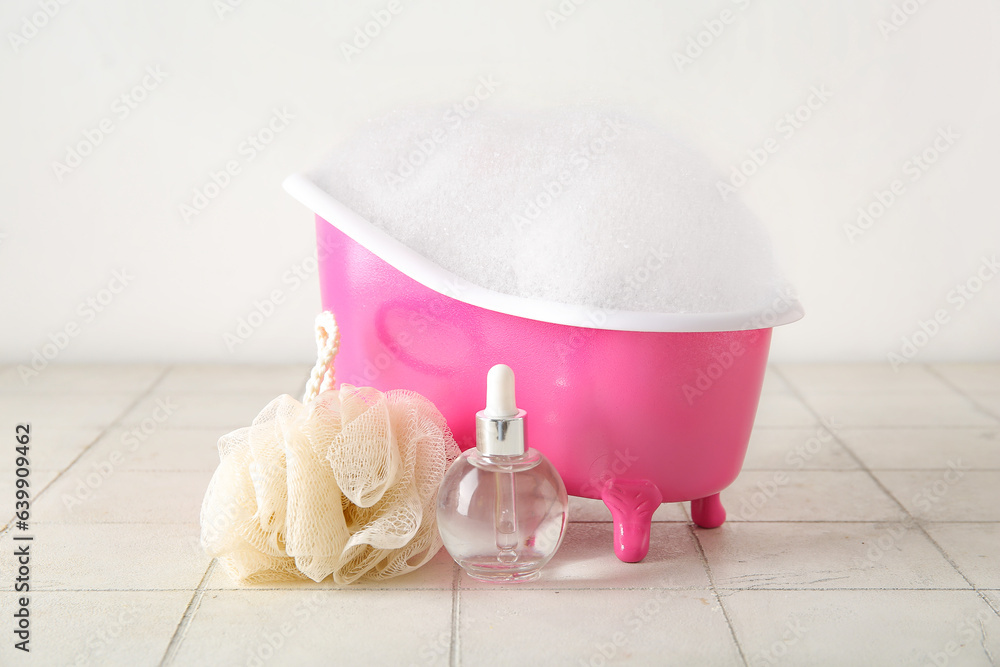 Small bathtub with foam, bottle of essential oil and sponge on light tile table