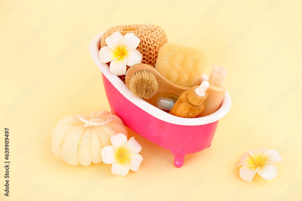 Small bathtub with different bath supplies and tropical flowers on color background