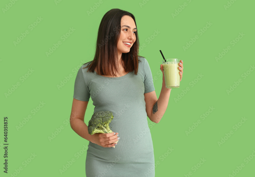 Young pregnant woman with glass of smoothie and broccoli on green background