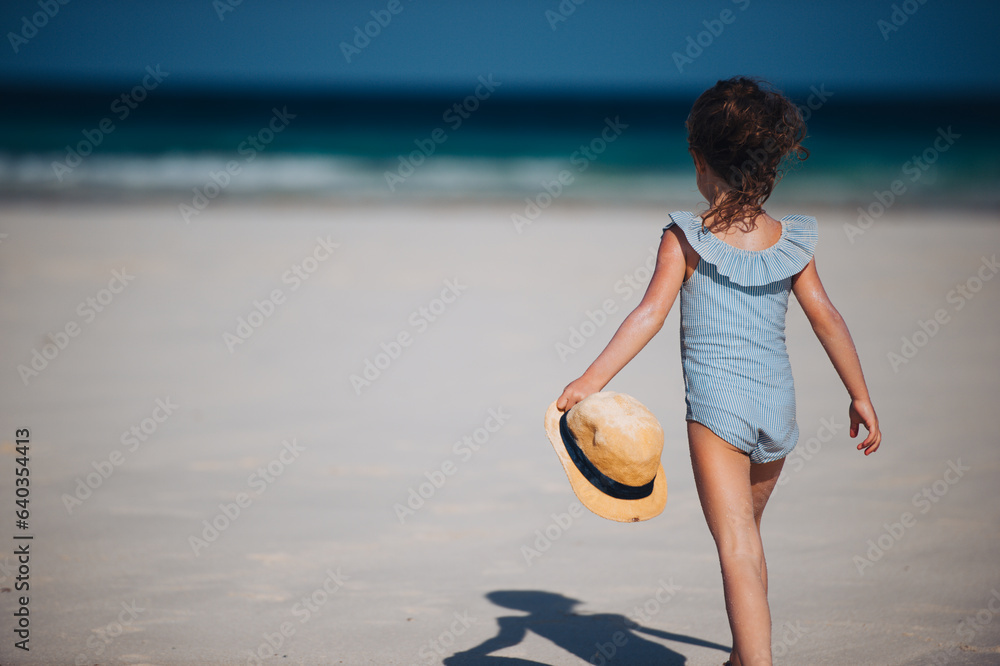 Rear view of a beautiful little girl walking on the beach in a swimsuit and a straw hat in hand.