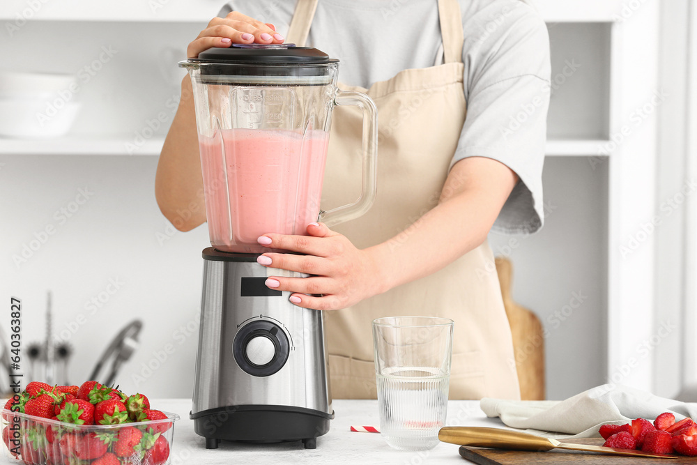 Woman preparing tasty strawberry smoothie with blender at table in kitchen