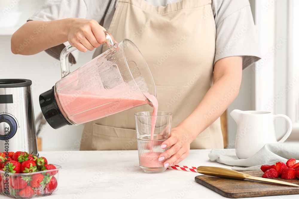 Woman pouring tasty strawberry smoothie from blender into glass at table in kitchen