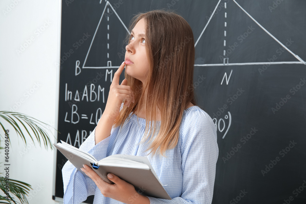 Pondering math teacher with book near blackboard in classroom