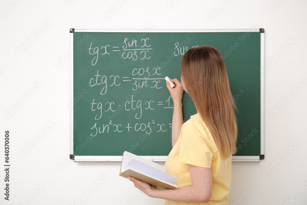 Young math teacher with book writing on blackboard in classroom