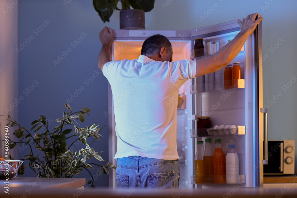 Hungry mature man near open fridge in kitchen at night, back view