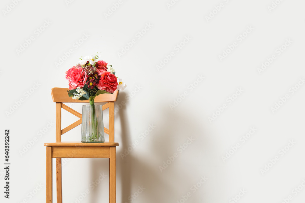 Vase with bouquet of beautiful artificial flowers on wooden chair near white wall