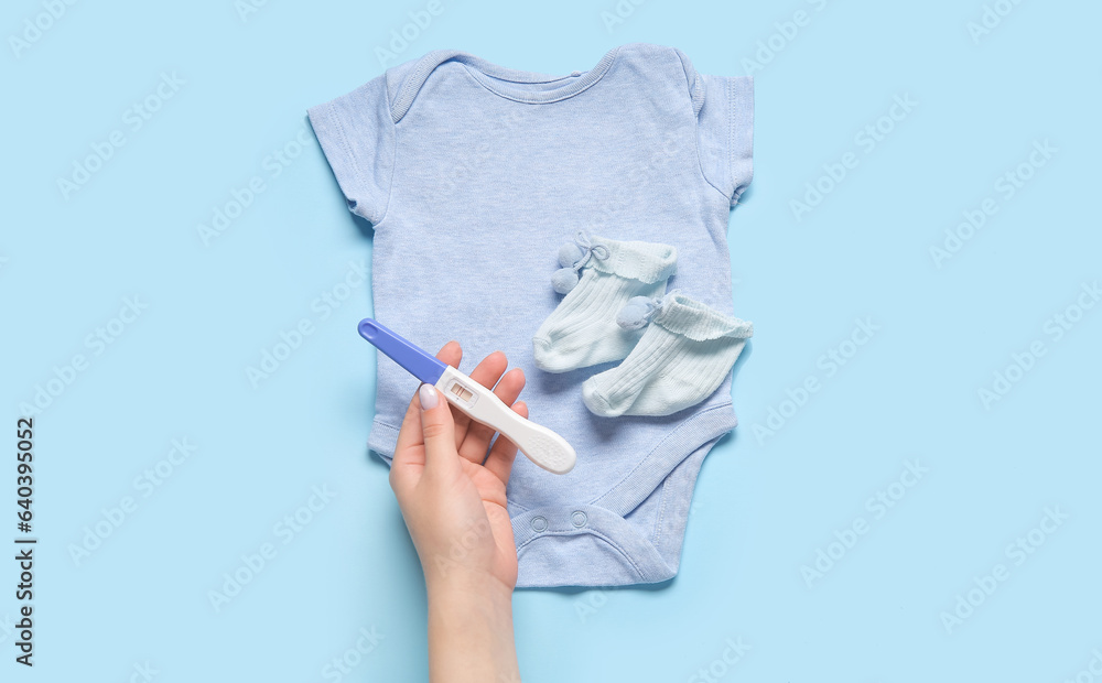 Female hand with pregnancy test and baby clothes on color background