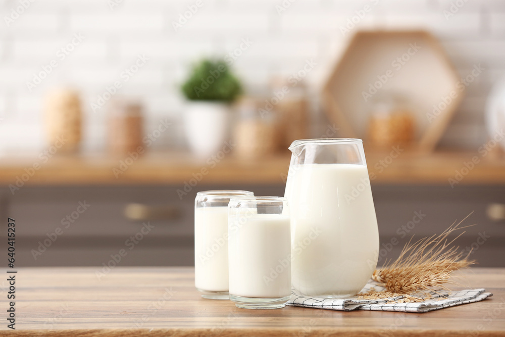 Glasses and jug of fresh milk with wheat ears on wooden table in kitchen