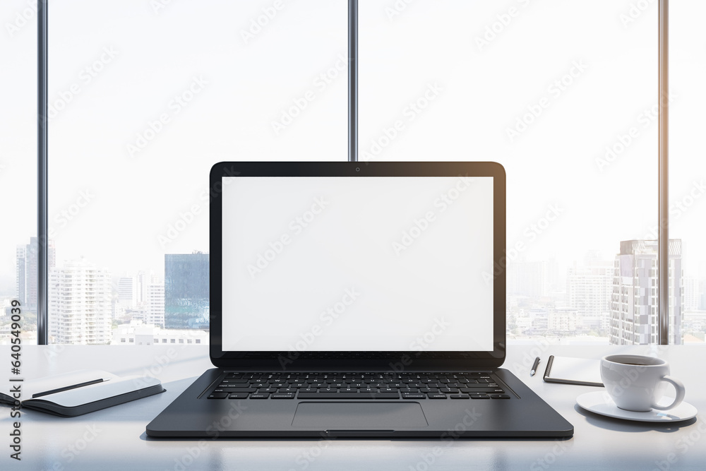 Close up of laptop with empty white mock up monitor, coffee cup and notepad placed on desk. Window w