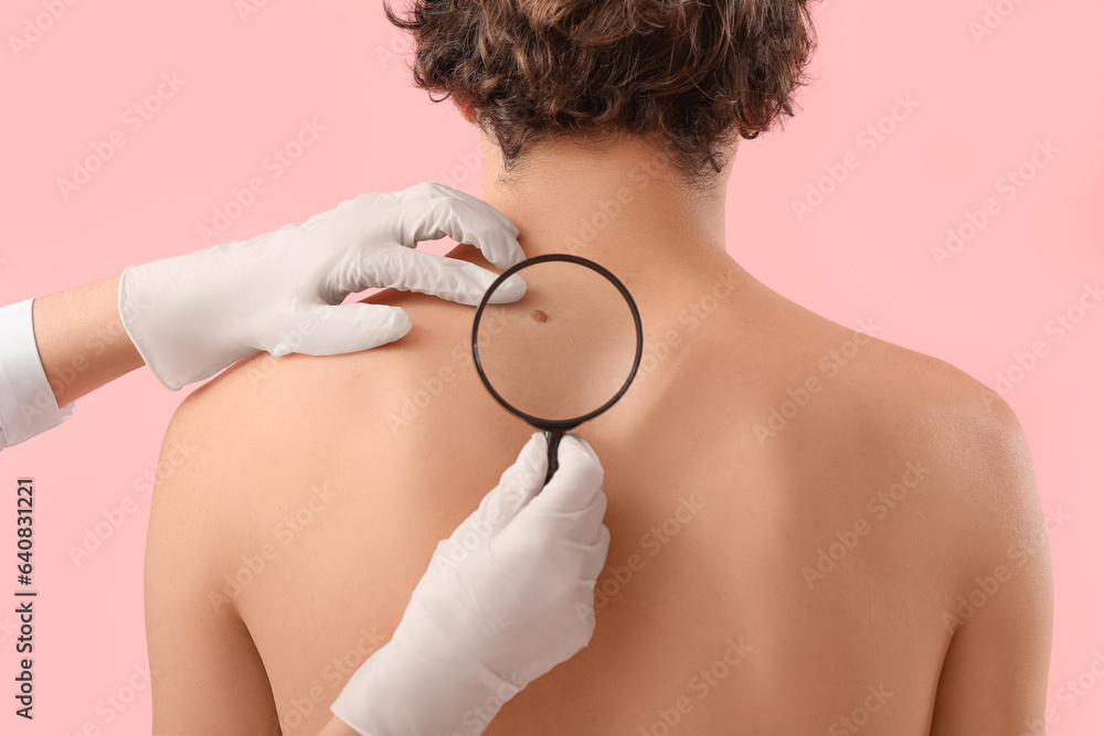 Dermatologist examining mole on young mans back with magnifier against pink background, closeup