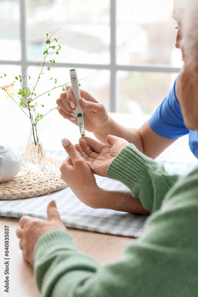 Young caregiver measuring sugar level of senior woman in kitchen, closeup