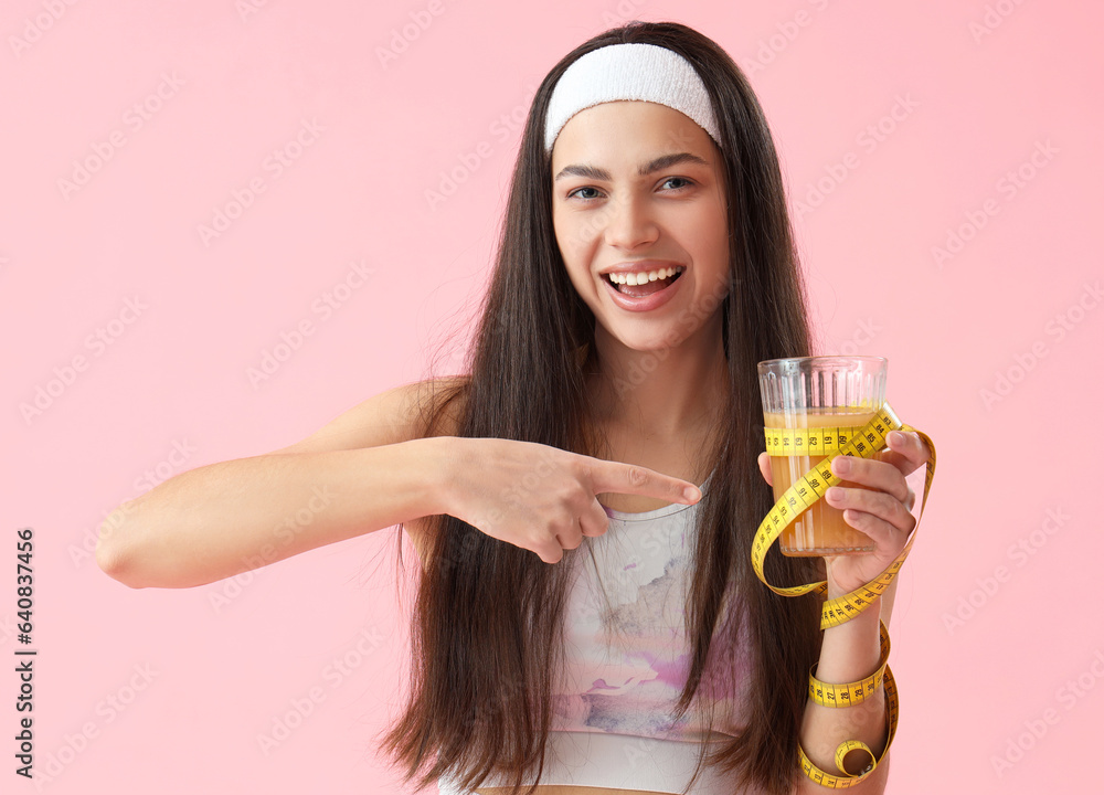 Sporty young woman pointing at glass of vegetable juice and tape measure on pink background