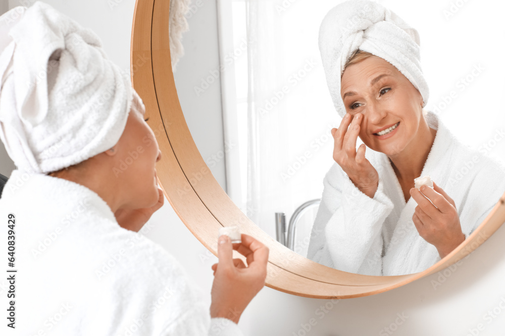 Mature woman applying under-eye cream near mirror in bathroom