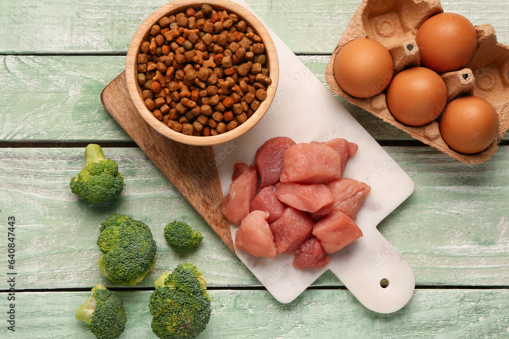 Bowl with dry pet food, raw meat, eggs and broccoli on color wooden background