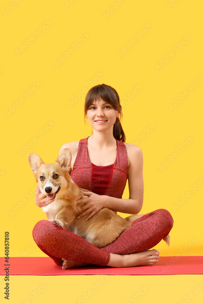 Sporty young woman sitting on yoga mat with cute Corgi dog against yellow background