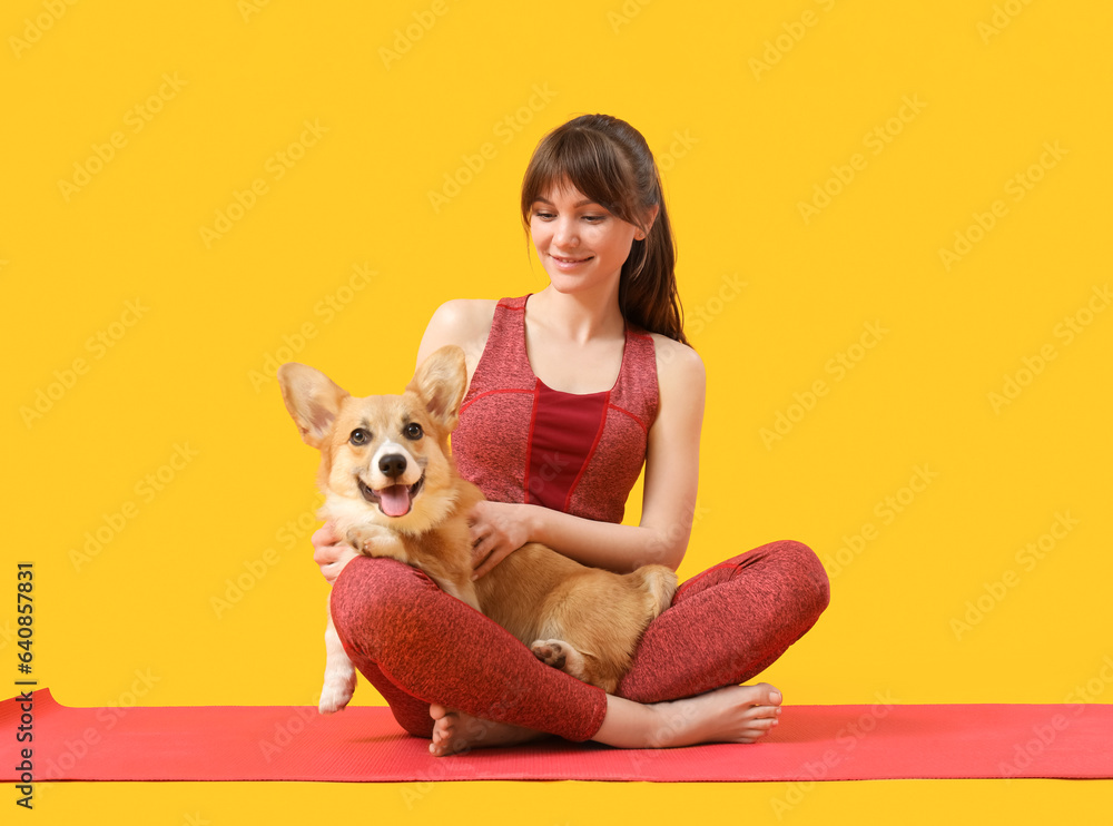 Sporty young woman sitting on yoga mat with cute Corgi dog against yellow background